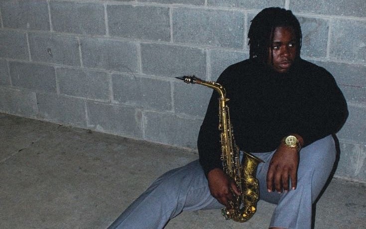 Brewster poses with his Saxophone, sitting against a concrete block wall