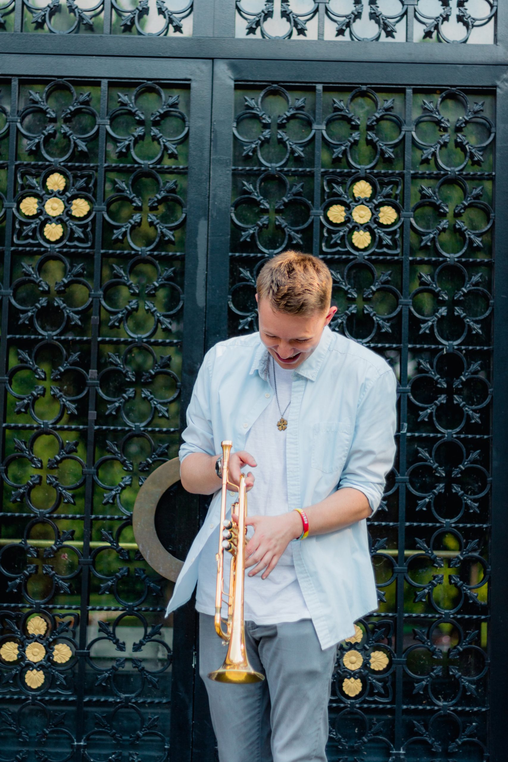 Lucas stands in front of a cast iron gate looking down and laughing, holding a trumpet