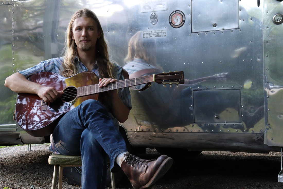 Pride sits on a vintage chair holding a guitar in front af an Airstream travel trailer.
