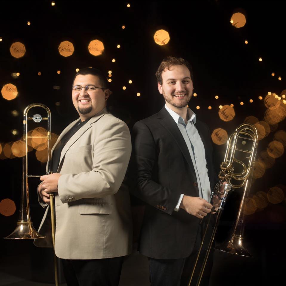 Altin Sencalar and Chris Glassman stand holding trombones against a backdrop of lights with a heavy bokeh lens effect